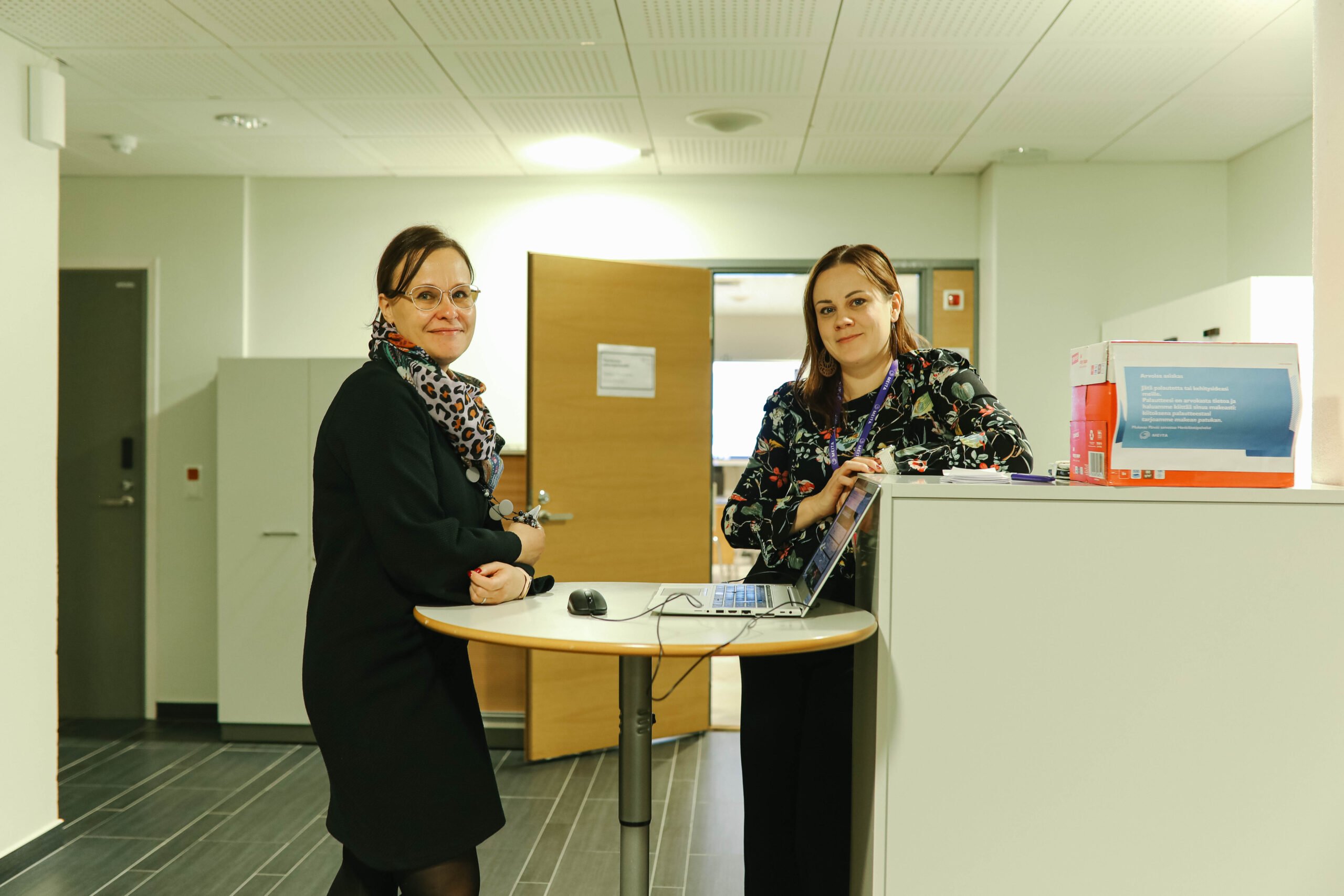 Tässä kuvassa on kaksi kameraan katsovaa meitalaista, joista toinen nojaa pöytään ja toinen kaappiin