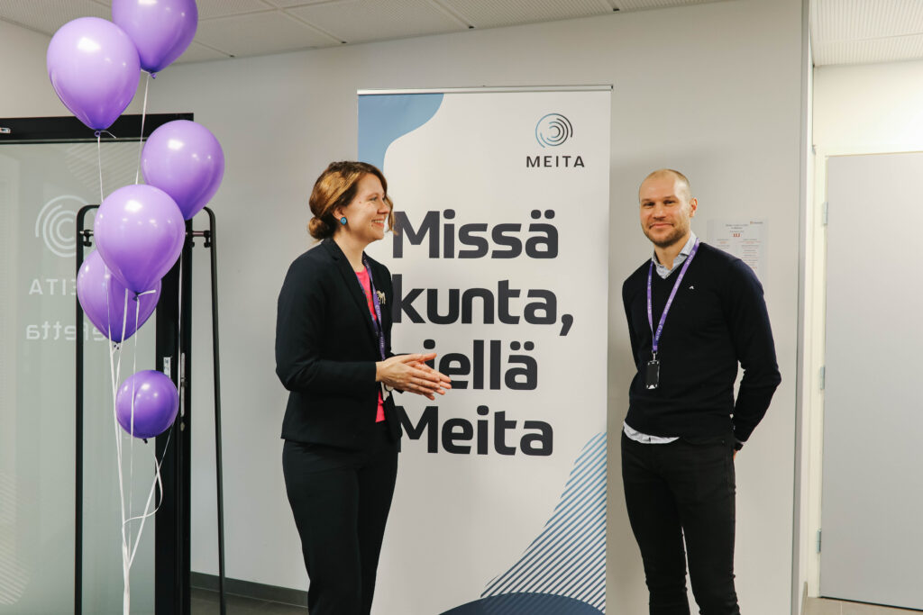 Tässä kuvassa kaksi meitalaista seisoo Meitan roll-upin edessä vierekkäin yhdessä nauraen ja kameraan katsoen ilmapallot vierellään